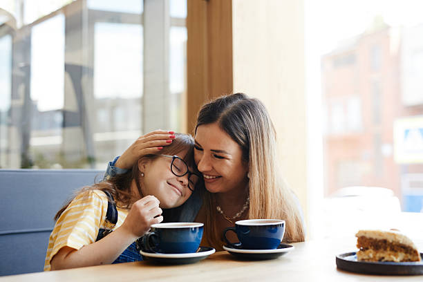 Portrait of loving mother embracing cute girl with down syndrome in cafe lit by sunlight, copy space