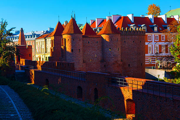 Historic brick fortifications with conical towers stand in the heart of Warsaw Old Town, bathed in warm sunlight. The medieval architecture contrasts beautifully with the colorful townhouses behind