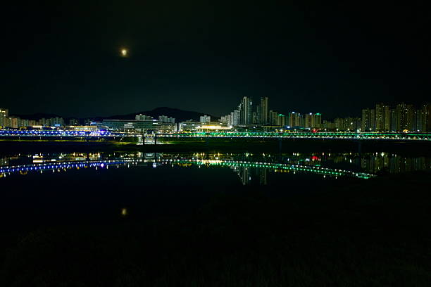 Sejong City, South Korea - June 12th 2025: The city skyline glows under a dim moon with lights stretching across a bridge and reflecting symmetrically on the calm surface of the Geum River.