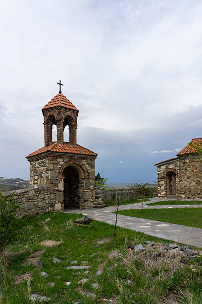 istock Medieval tall bell tower with brown tiled roof. Low stone wall approaches from both sides. Stone path leads to the entrance. Green grass, blue sky 2222200322