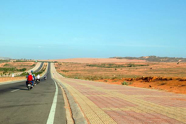Mui Ne, Vietnam - March 25, 2018: Motorbikes Ride Through Coastal Desert Landscape Under Blue Sky.