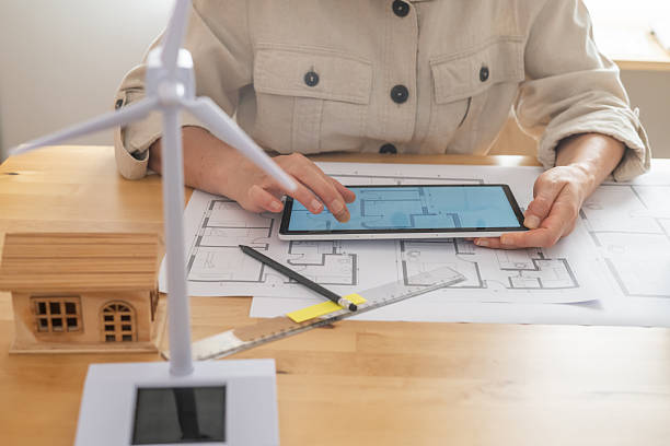 istock Architect is using a tablet to design a sustainable and ecological house powered by renewable energy, with blueprints, a miniature wind turbine, and a miniature house model on the desk 2222204459