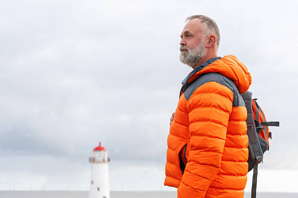 A man in a bright orange jacket gazes thoughtfully towards the horizon near a lighthouse. The weather is overcast, creating a moody atmosphere.