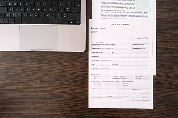 Flat lay of collection of paperwork, including an application form and contract, is neatly arranged on a wooden desk alongside a laptop. Workspace highlights productivity in home office, top view
