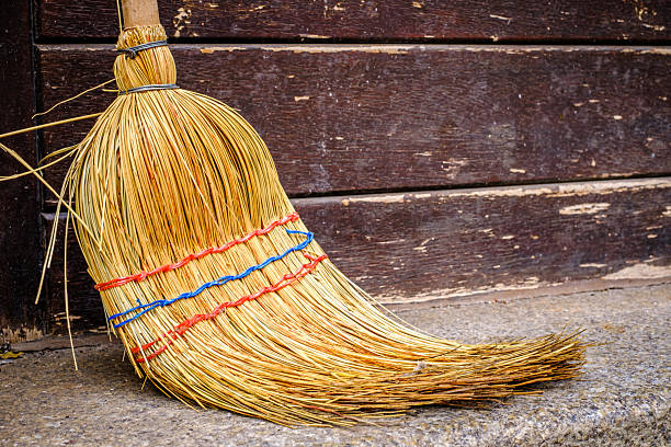 A straw broom leans against a wooden shelf in a rustic environment, illuminated by warm afternoon light, highlighting the simplicity and craftsmanship of the scene.