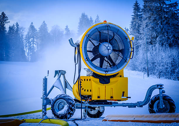 A snowmaking machine is actively producing artificial snow at a ski resort. The surrounding area features evergreen trees and a snowy landscape under a blue sky.