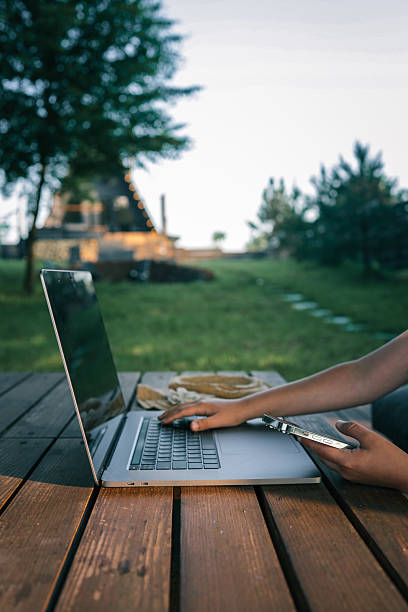 istock Person using a laptop and holding a smartphone while sitting at a wooden table outdoors, with a cozy cabin in the background and green trees surrounding the area 2222251384