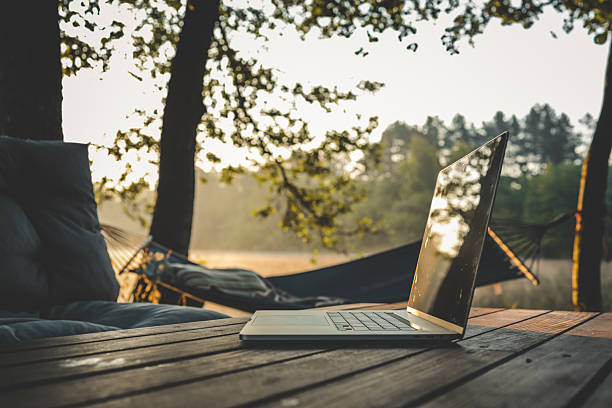Laptop placed on a wooden table in a tranquil forest setting with a hammock and soft morning light, ideal for remote work or digital nomad lifestyle.