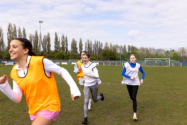 A three-quarter length shot of a group of girls in a girls' under-15s football team wearing sports clothing, football trainers and sports bibs. They are warming up together, running laps before they begin football practice. Located in the North East of England.Videos are available for this scenario.