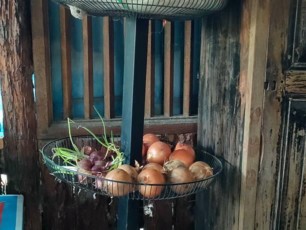 a wire hanging basket filled with red and yellow onions, some of which have sprouted green shoots, indicating freshness and potential for regrowth. The basket is part of a tiered storage rack standing against a rustic wooden wall, adding to the traditional, countryside kitchen vibe. The surrounding textures—aged wood, bamboo window bars, and soft lighting—create a warm and nostalgic atmosphere, capturing the charm of simple rural living.