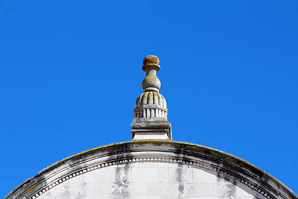 Ornate stone finial with weathered details sits atop a curved building cornice, highlighted against a clear blue sky, showing classical architectural style and aged surface