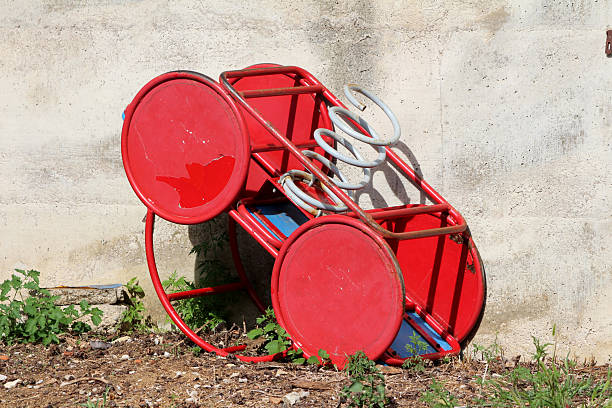 Red metal playground spring rider with circular seats and white coil spring lies overturned on soil and weeds, leaning against a weathered concrete wall in sunlight