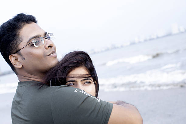 Young Indian Couple Embracing on a Picturesque Beach in Mumbai, India, Gazing into the Distance while the Woman's Hair Dances in the Gentle Sea Breeze, Radiating Joy and Intimacy