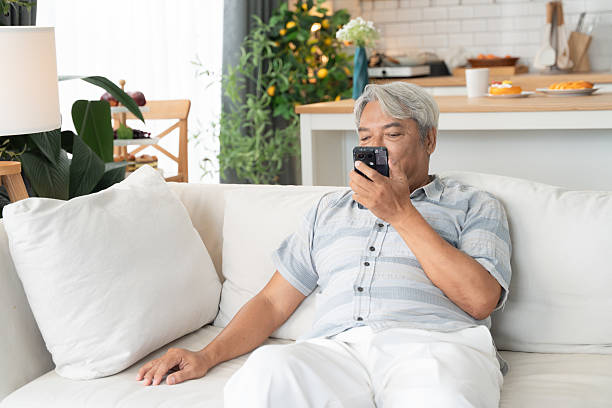istock Senior Asian man relaxing on sofa at home while using smartphone, communicating with voice message. Modern lifestyle, elderly technology usage, and comfortable living concept. 2222284900