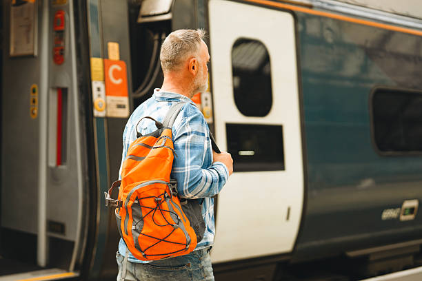 A man stands on a train station platform, wearing a plaid shirt and holding a coffee. His bright orange backpack contrasts with the train behind him as he waits.