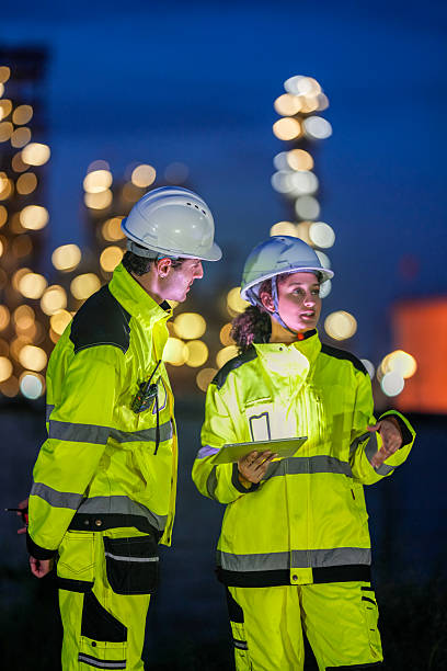 istock Two engineers in reflective safety gear review plans on a tablet during a night shift at an industrial site, with blurred factory lights glowing in the background. 2222293696