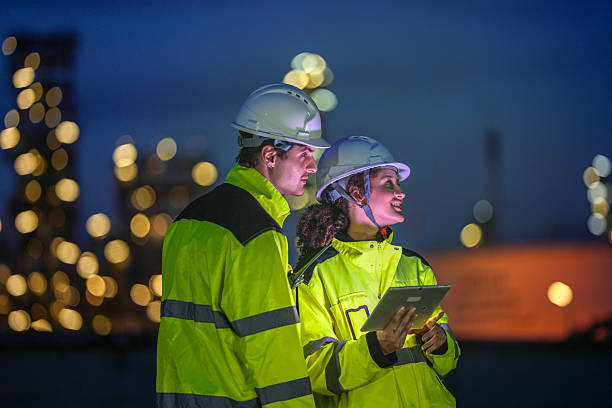 istock Two engineers in reflective safety gear review plans on a tablet during a night shift at an industrial site, with blurred factory lights glowing in the background. 2222293721