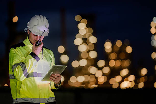 istock Male engineer in a reflective safety jacket and hard hat communicates via radio while checking information on a tablet, with an industrial facility glowing in the night background. 2222293742