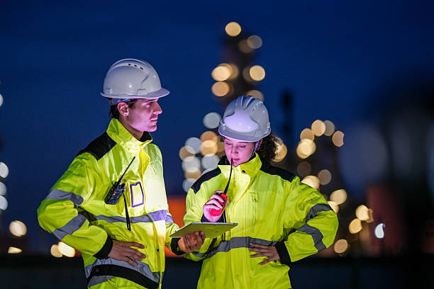 istock Two engineers in high-visibility jackets and safety helmets work during the night shift at an industrial facility, reviewing data on a tablet and coordinating via walkie-talkie. 2222293755