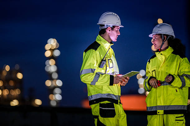 istock Two engineers in high-visibility jackets and safety helmets work during the night shift at an industrial facility, reviewing data on a tablet and coordinating via walkie-talkie. 2222293791