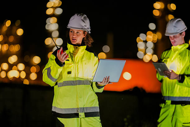 istock Female engineer in high visibility jacket uses a radio while analyzing data on a laptop during a night inspection at an industrial site, with blurred lights in the background. 2222294944