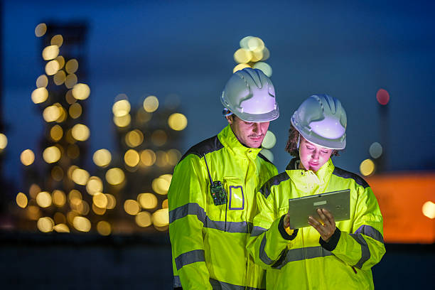istock Two engineers in reflective safety gear review plans on a tablet during a night shift at an industrial site, with blurred factory lights glowing in the background. 2222295144