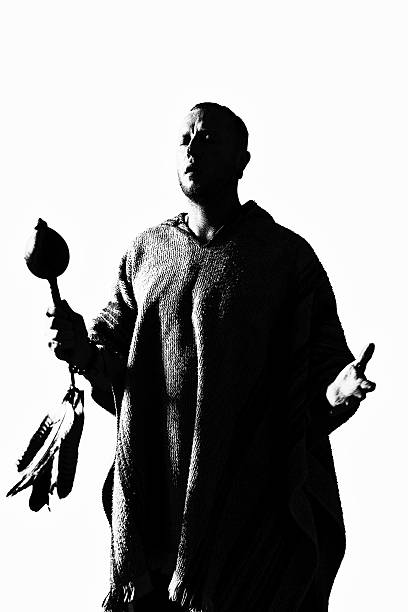 Black and white high-contrast image of a man wearing a textured poncho, holding a ceremonial rattle with feathers. His eyes are closed in deep focus, with one hand raised in an expressive, open gesture. The minimalist background and shadowed figure evoke themes of ancient wisdom, healing, and spiritual ritual. A modern interpretation of timeless shamanic practice.