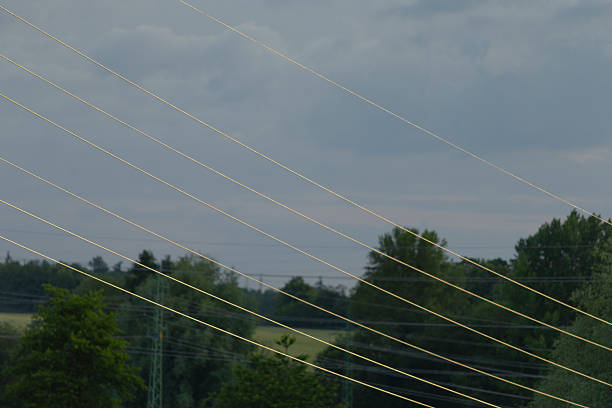 rural landscape at sunset, with a towering electricity pylon standing prominently in the middle of a lush, green field. Power lines stretch across the frame, disappearing into the distance, highlighting the intersection of nature and technology. Rolling hills covered in dense trees surround the area, with soft orange and pink hues blending into the blue sky as daylight fades. The serene natural setting contrasts with the industrial presence of the power infrastructure, making it an ideal image for themes related to energy, nature, or rural development