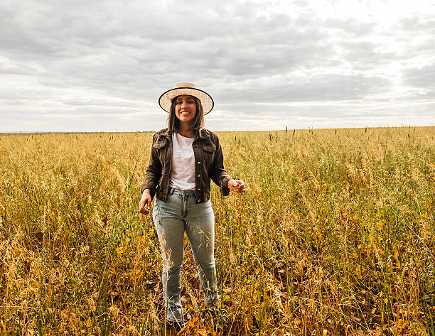 A woman in her 30s walks alone through a rural agricultural field filled with tall grasses or grains, possibly oats or wheat. She wears a wide-brimmed hat, casual clothing, and has light skin, conveying a connection to nature and rural life. The photo was taken during the daytime under a bright but cloudy sky, indicating overcast weather. The scene reflects simplicity, countryside lifestyle, and sustainable farming.