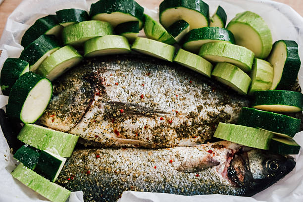 Two whole fish seasoned with herbs sit alongside fresh zucchini slices in a baking dish prepared for a delicious meal.