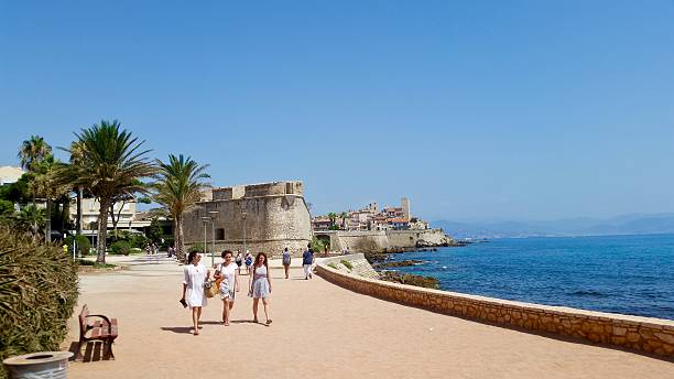Antibes, France, august 02, 2017. a sunny day on the coastal walkway, with people strolling along the path next to the sparkling Mediterranean Sea. In the background, the historic town of Antibes with its fortified walls and a prominent clock tower is visible, nestled on the rocky coastline. Palm trees and lush vegetation line the promenade