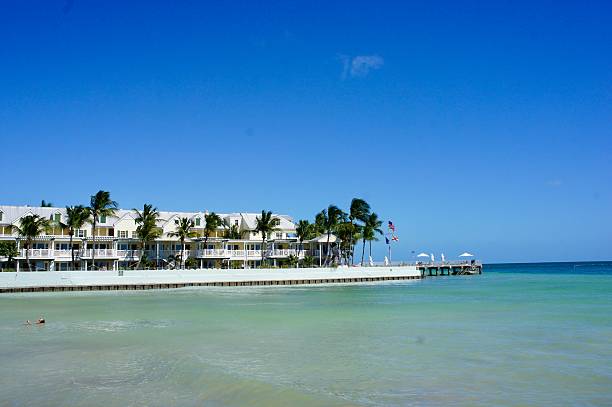 Stunning beachfront residence in Key West, Florida, featuring pastel facades, private balconies, and palm trees swaying in the sun. The turquoise pool and pier adorned with American flags create an exclusive vacation atmosphere, facing a calm, clear sea. The cloudless blue sky and crystalline water evoke the tropical lifestyle and unique charm of this iconic destination.