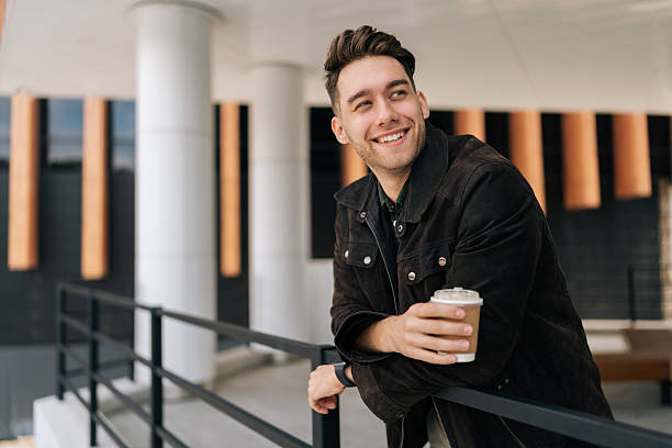 istock Handsome positive young man relaxing during coffee break, leaning on railing standing by modern office building. Happy bearded male in casual attire smiling looking away. 2222337542