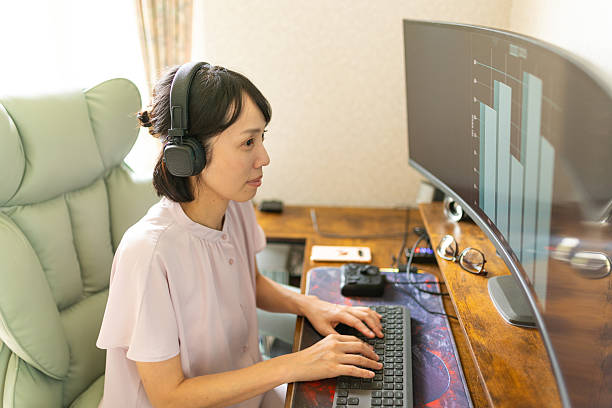 A Japanese woman at her home computer desk, working on a graph with an ultra-wide curved monitor.