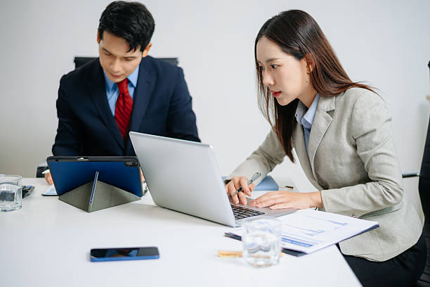 istock Two Asian business professionals in formal suits discussing a project at modern office desk with laptop and documents. 2222346547
