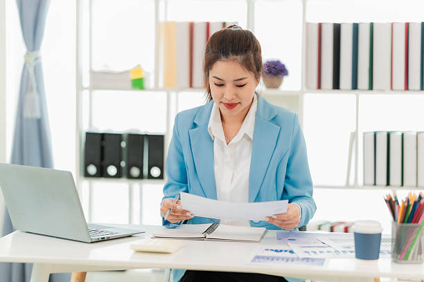 istock Successful Asian professional businesswoman in suit sitting smiling, reading, analyzing data on documents and laptop, working alone in corporate office. 2222348688