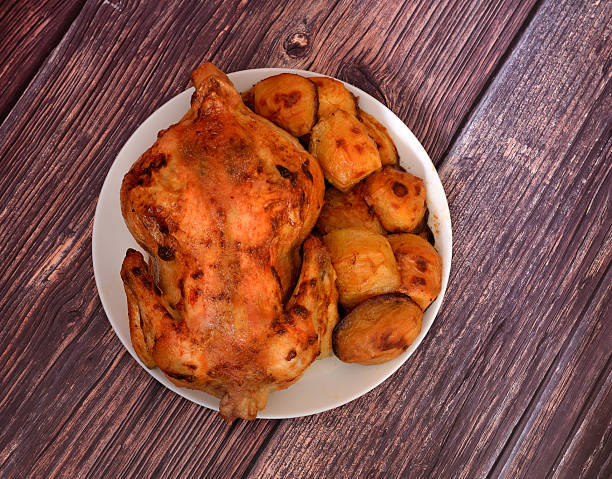 Whole baked domestic chicken with new potatoes on a large round plate on a wooden table.