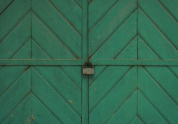 A close-up view of a textured green wooden gate secured with a sturdy metal padlock. The geometric wooden patterns add an intricate and symmetrical detail to the rustic design.