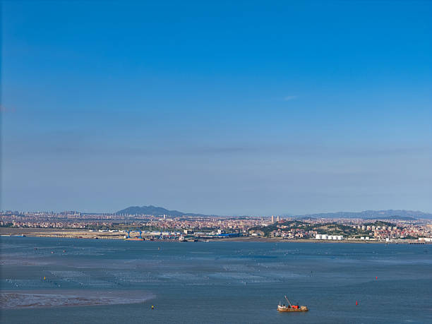 A serene coastal city view featuring calm waters and distant urban landscape. Visual style is naturalistic, capturing tranquility and potential for travel or real estate promotion.