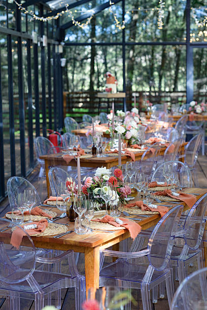 An elegant wedding reception showcasing beautifully decorated tables with floral centerpieces and glass tableware, set under a glass pavilion surrounded by lush greenery. The rustic design combined with modern transparent chairs provides a charming atmosphere, perfect for a celebration of love and togetherness.