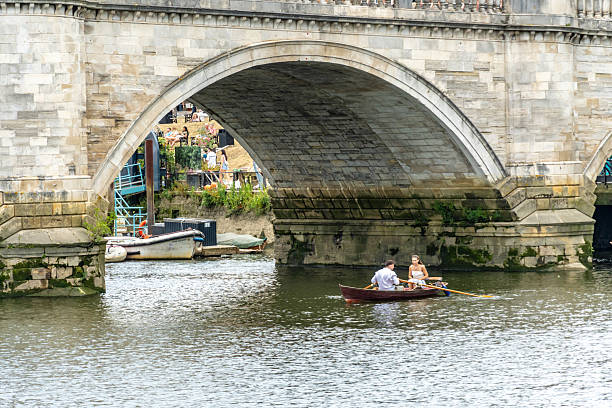 Aerial view of boats on a river beneath an arched stone bridge, surrounded by buildings and trees on a sunny day  in Knaresborough, UK.