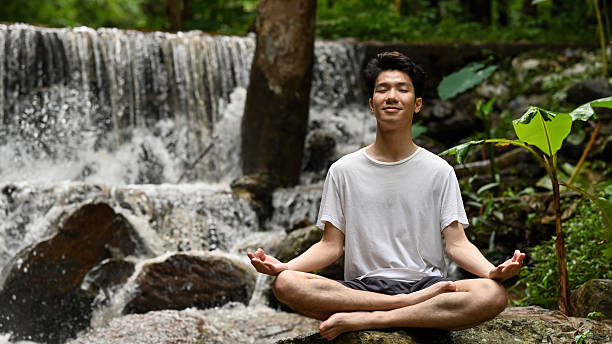 istock Young man sits peacefully in a meditative pose by a waterfall in the forest. Eyes closed, calm expression, connecting with nature in a serene 2222395014