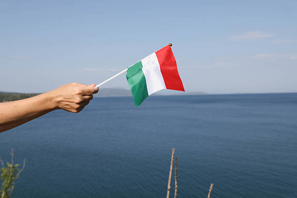 A senior woman's hand holding the Hungarian flag beside the sea, symbolizing national pride, identity, and a connection to heritage. Captured in a serene coastal setting, this scene evokes themes of patriotism, summer travel, and European culture. Suitable for topics such as tourism in Hungary, diaspora, or Mediterranean vacations with a personal and emotional touch.