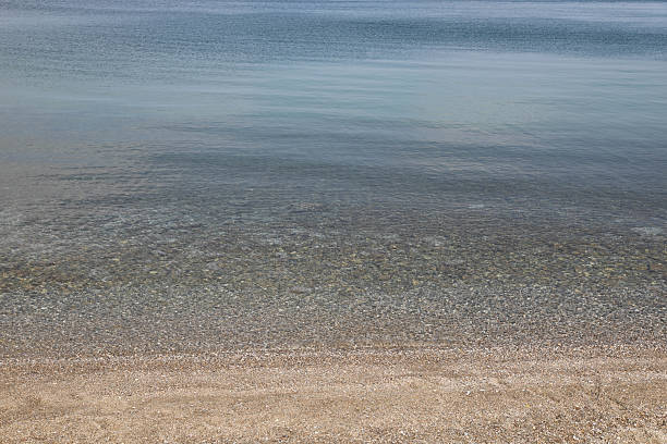 Tranquil seascape with crystal-clear water and a pebble beach. The calm waves and gradient tones from sandy shore to deeper blue create a peaceful, minimalist composition. Ideal for backgrounds, travel themes, and relaxation concepts.