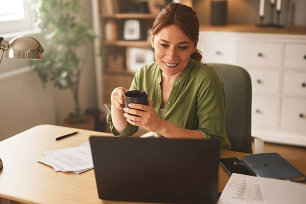 istock Woman working from home drinking coffee and smiling at laptop 2222401738