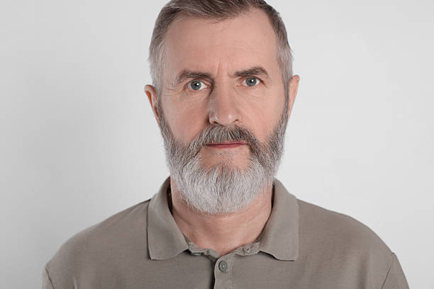 An elderly 50-55 year old man with a beard wearing a black hoodie looks into the camera against a light colored background