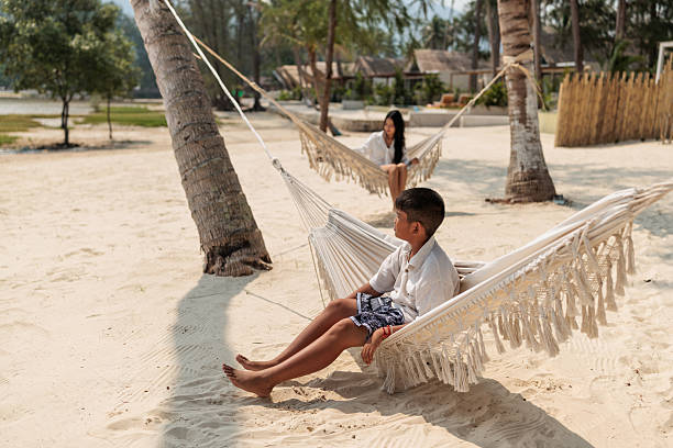 A brother and sister lounge comfortably on hammocks tied to palm trees, enjoying the gentle sea breeze and the soothing sounds of the beach.