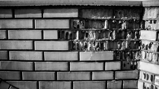 Close-up of an old brick wall with severely damaged facing tiles, revealing the internal structure beneath. Captured in black and white, the image emphasizes texture, decay, and urban wear.
