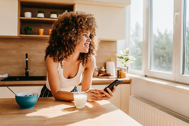 Smiling young woman using smartphone and having breakfast in kitchen