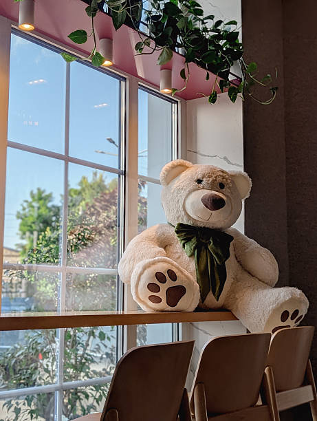 Giant teddy bear resting on a windowsill in a warmly lit cafe. Interior with chairs, tables, indoor plants, and a sunny outdoor view.
